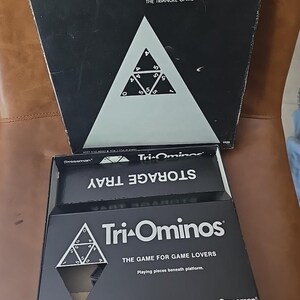 May include: A black Tri-Ominos board game box with a white triangle design and the text "Tri-Ominos THE TRIANGLE GAME". The storage tray is also black with white text. The game is for game lovers.