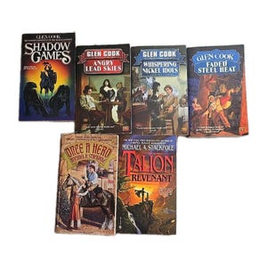 May include: A collection of fantasy novels by Glen Cook and Michael A. Stackpole. The books feature colorful covers with titles like "Shadow Games," "Angry Lead Skies," and "Talon: Revenant." The books are arranged on a white surface.
