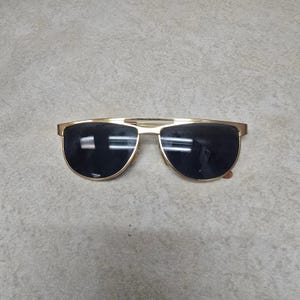 May include: Gold-tone sunglasses with dark lenses. The sunglasses have a double bridge and a classic aviator style. The lenses are a dark, reflective color, and the frames have a shiny gold finish. A stylish accessory for eye protection.