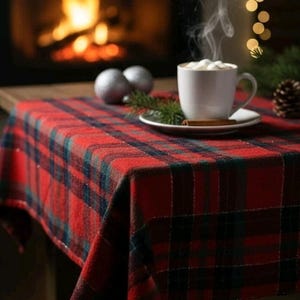 May include: A red and dark green plaid tablecloth draped over a table. A white ceramic mug filled with a hot beverage topped with white marshmallows sits on a matching saucer, accompanied by a cinnamon stick and a pine sprig.