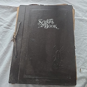 May include: A vintage scrapbook with a dark brown cover. The cover features the words "Scrap Book" in an ornate font, along with a windmill illustration. A string is tied around the spine to keep the pages together.