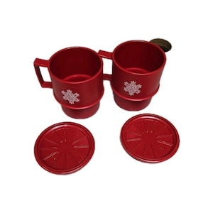 May include: Two red mugs with handles and snowflake designs, each with a matching coaster. The mugs have a stepped base and are made of a solid red material. The coasters are round with a radial pattern.