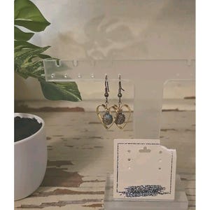 May include: A pair of heart-shaped earrings with a gold-colored frame and a textured, multi-colored center. The earrings are displayed on a clear acrylic stand with a white earring display card.