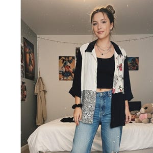 May include: A color-blocked button-down shirt with black, white, and patterned panels. The shirt is worn over a black top and paired with blue jeans. The outfit is completed with a black scrunchie and layered necklaces.