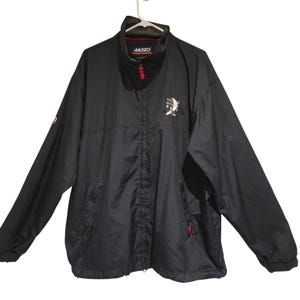 May include: Black Musto jacket with a high collar and a red zipper. The jacket features the Musto logo on the collar and a white and black graphic on the chest. The jacket has two front pockets with red accents and a small logo on the left sleeve.