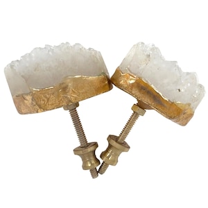 May include: Two cabinet knobs featuring raw, white crystalline druzy stone tops with irregular, jagged edges. Each stone is encased in a thick, textured gold-toned metal border. A threaded metal bolt and a decorative brass-colored base are attached.