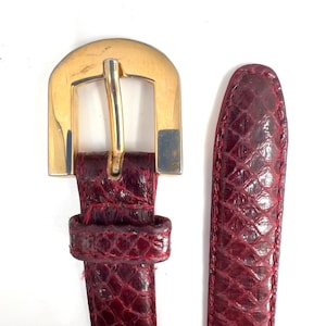 Puede incluir: Un cinturón de color burdeos intenso con un patrón texturizado similar a escamas y una hebilla de metal en tono dorado pulido. La correa del cinturón tiene costuras a juego en los bordes y la hebilla presenta un diseño de marco semicircular.