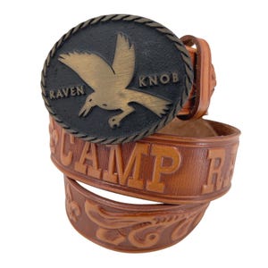Könnte beinhalten: Ein brauner Ledergürtel mit einer großen, kreisförmigen Schnalle. Die Schnalle zeigt ein bronzenes Raben-Design auf schwarzem Hintergrund, mit den Worten "Raven Knob" am Rand. Der Gürtel ist mit dem Wort "Camp R" und dekorativen floralen Mustern geprägt.