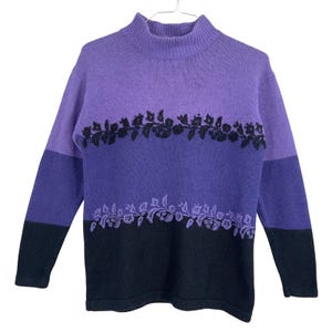 May include: A long-sleeve mock neck sweater featuring a color-blocked design in shades of light purple, medium purple, and black. The sweater includes two horizontal bands of floral embroidery, with black flowers on the upper chest and purple flowers below.