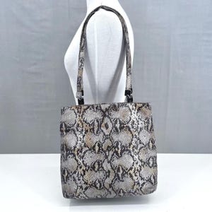 May include: A square-shaped handbag with a snakeskin pattern in shades of gray, brown, and beige. The bag has two handles and black hardware. The bag is displayed on a white surface.