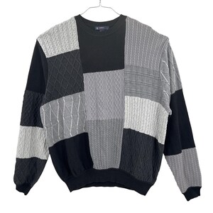 Pode incluir: Um suéter patchwork em tons de preto, cinza e branco, com várias texturas de malha. O suéter tem gola redonda e mangas compridas, pendurado em um cabide de metal sobre um fundo branco.