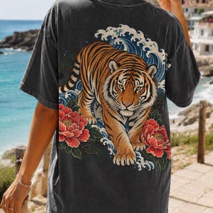 May include: A charcoal gray short-sleeve t-shirt featuring a large graphic on the back. The design displays a tiger walking forward, surrounded by stylized blue ocean waves and vibrant red peony flowers with green leaves on a dark background.