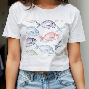 Peut inclure: T-shirt blanc orné d'un motif de poissons dessinés en bleu, vert et rouge. Les poissons sont détaillés avec des lignes texturées. Le t-shirt est de style court, une tendance actuelle.