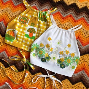 May include: Two halter tops with adjustable straps. One top is yellow and white with a gingham pattern and mushroom and sailboat designs. The other top is white with a floral pattern.