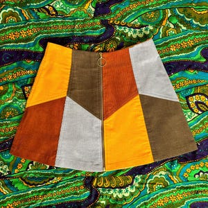 May include: A patchwork mini skirt made of corduroy in shades of orange, brown, and gray. The skirt has a zipper down the front and a geometric pattern.