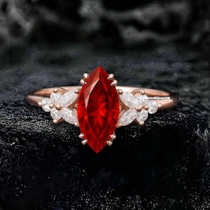 May include: Rose gold ring featuring a large, marquise-cut red gemstone as the centerpiece. The ring is accented with smaller, white gemstones in a floral design on either side of the main stone. The ring is set against a dark background.