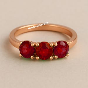 Pode incluir: Um anel de ouro rosa com três pedras preciosas vermelhas vibrantes. O anel tem uma banda lisa e polida e as pedras são fixadas com garras. O texto "TimelessBijouxUK" é visível no anel.