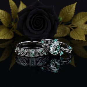 May include: Two silver rings with green gemstones. One ring features an oval-cut stone and leaf accents. The other ring has a rectangular green stone and a detailed band. A black rose and gold leaves are in the background.