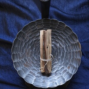 Könnte beinhalten: Eine dunkelgraue, kreisförmige Metallschale mit gewelltem Rand und texturierter, gehämmerter Oberfläche. Ein einzelnes Stück hellbraunes, grob behauenes Holz ist mit einer dünnen, weißen Schnur zusammengebunden und liegt in der Mitte der Schale.