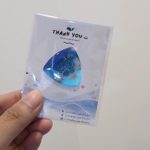 May include: A triangular guitar pick made of blue and white resin, featuring a small embedded starfish and bubble-like textures. It is packaged on a white card with the text THANK YOU, have a good day!! and social media handles for LittleBeluga.Studio.