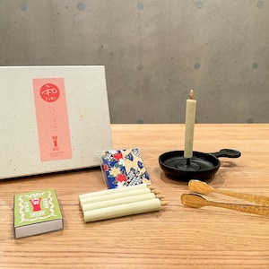 以下が含まれることがあります： ピンクのラベルが付いた白い箱、小さな緑色のマッチ箱、火のついたろうそくが入った黒い燭台、装飾的なトングなど、日本のスタイルのアイテムのコレクション。 また、いくつかの消灯したろうそく、小さな模様のカードも含まれています。