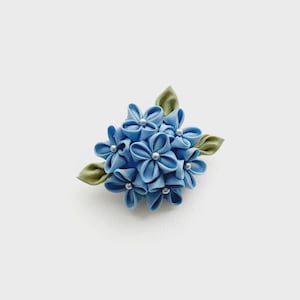 以下が含まれることがあります： 複数の水色の花びらを房状に配置した布製のフラワーブローチ。各花の中心には小さな銀色のビーズがあしらわれ、側面には尖ったセージグリーンの布製の葉が数枚添えられています。