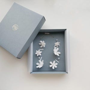 Puede incluir: Una caja de regalo cuadrada gris con una tapa que presenta un pequeño logotipo circular, que contiene un delicado conjunto de accesorios para el cabello. Incluye varias flores de tela blanca con perlas en el centro sobre un hilo dorado.