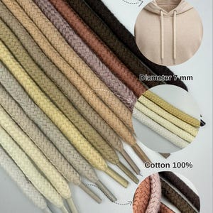Puede incluir: Una colección de cordones trenzados de algodón para sudaderas con capucha en varios colores neutros, como beige, marrón y gris. La imagen muestra los cordones con un diámetro de 7 mm, con los textos "Great for hoodies" y "Cotton 100%" visibles.