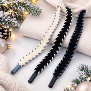 May include: A cream-colored hoodie featuring three thick, flat drawstrings with braided edges. One drawstring is cream with light gray stitching, while the other two are black with white and light gray stitching. Each drawstring has a solid black tip.