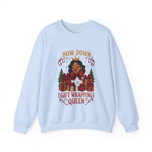 Può includere: Felpa azzurra con la scritta "BOW DOWN TO GIFT WRAPPING QUEEN". La grafica presenta una donna incoronata, regali incartati e alberi di Natale. Il testo è in oro e rosso.