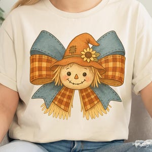 May include: Cream-colored t-shirt featuring a whimsical scarecrow design. The scarecrow has a straw hat with a sunflower, a plaid scarf, and a blue bow. The design is in shades of orange, yellow, and blue.