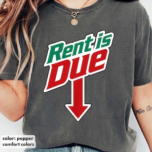 May include: A pepper-colored short-sleeve t-shirt featuring the text Rent is DUE in a stylized graphic. The word Rent is in green, while the word DUE is in red with a large red downward-pointing arrow underneath, all outlined in white.