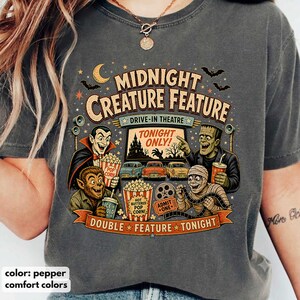 Puede incluir: Una camiseta color pimienta con un estampado gráfico que incluye el texto MIDNIGHT CREATURE FEATURE DRIVE-IN THEATRE TONIGHT ONLY. El diseño muestra ilustraciones de un vampiro, un hombre lobo, una momia, palomitas, coches y rollos de película.