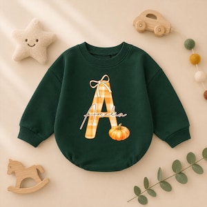 Puede incluir: Una sudadera de manga larga de color verde oscuro con una letra A grande con un patrón de cuadros naranja y blanco, un pequeño lazo en la parte superior, el nombre Amelia escrito en cursiva sobre la letra y un pequeño gráfico de calabaza.
