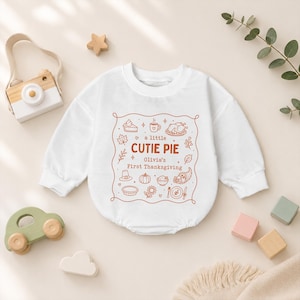 Pode incluir: Um body de bebé branco de manga comprida com uma impressão gráfica cor de ferrugem. O design inclui o texto 'a little CUTIE PIE' e 'Olivia's First Thanksgiving' rodeado por ilustrações de um peru, tarte, abóbora e folhas de outono.