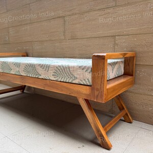 Puede incluir: Un banco de madera con un cojín rectangular de color claro que presenta un patrón repetitivo de hojas verdes. El banco tiene un acabado de madera natural, reposabrazos en ambos extremos y patas inclinadas que sirven de soporte.