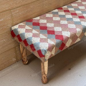 Peut inclure: Un banc rectangulaire doté d'un dessus tissé avec un motif en losanges répétitif dans des tons de rouge, gris et beige. Le banc est soutenu par quatre pieds en bois tourné avec une finition en grain de bois brun clair naturel et une texture lisse.