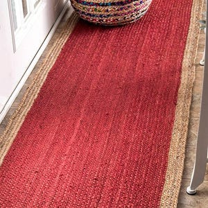 Puede incluir: Una alfombra rectangular de yute rojo con un borde de yute natural. Un puf redondo y multicolor se encuentra sobre la alfombra. La alfombra está hecha de fibras tejidas y está colocada sobre un suelo embaldosado.