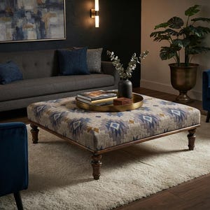 Pode incluir: Um grande otomano quadrado com tecido estampado em tons de azul, bege e castanho, apoiado em quatro pernas de madeira escura torneadas. Uma bandeja redonda dourada repousa no topo, contendo um vaso preto com eucalipto e livros.