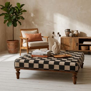 Peut inclure: Un pouf carré présentant un motif à damier noir et beige sur sa surface supérieure, soutenu par quatre pieds en bois brun foncé au design tourné. Le pouf est placé dans une pièce avec un sol clair et des meubles en bois.