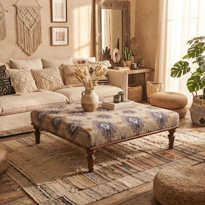 Può includere: Un grande pouf quadrato rivestito in tessuto con motivi geometrici blu, gialli e bianchi, sostenuto da quattro gambe in legno tornito marrone scuro. Il pouf è posizionato in un soggiorno con un divano beige e cuscini da pavimento intrecciati.