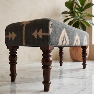 May include: A rectangular upholstered bench featuring a dark charcoal gray fabric with a repeating beige arrow and leaf-like pattern. The bench is supported by four dark brown turned wooden legs with a smooth, polished finish and rounded details.