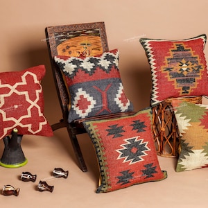 Puede incluir: Una variedad de cojines decorativos con patrones geométricos en rojo, negro, blanco y verde. Los cojines están colocados sobre una silla de madera y una cesta tejida, con pequeños objetos decorativos en primer plano.