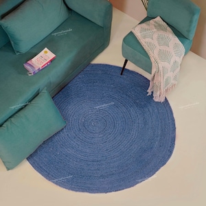 May include: A teal sofa and armchair with a blue round rug. A throw blanket with a white and green pattern is draped over the armchair. A small book is on the sofa. The rug is made of woven material.