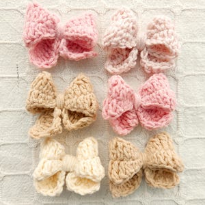 May include: Six handmade crocheted bows in pink, beige and cream. The bows are arranged in two rows of three.