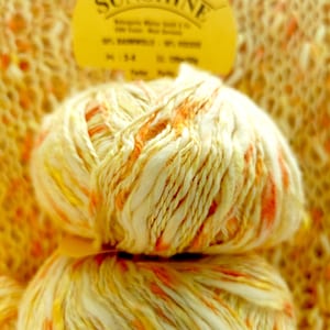 Puede incluir: Dos ovillos de hilo amarillo, blanco y naranja. El hilo está etiquetado como "Sunshine" y está hecho de 100% bambú.