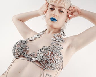May include: A silver metal bra with a unique, open design. The bra features intricate wirework and chain details. The person in the image has blue lipstick and short blonde hair. The overall aesthetic is modern and artistic.