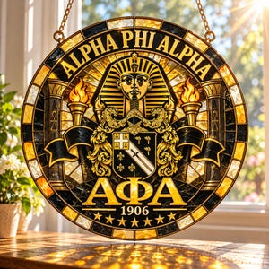 May include: A circular stained glass suncatcher featuring the Alpha Phi Alpha fraternity crest. The design includes a golden pharaoh head, two flaming torches, a shield with a sword and fleur-de-lis, the text ALPHA PHI ALPHA, AΦA, and 1906.
