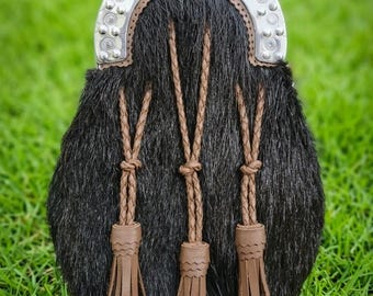 May include: A black fur sporran featuring a silver-toned metal cantle with embossed circular detailing. The front is adorned with three brown braided leather tassels that hang from matching leather cords, creating a textured and functional accessory.