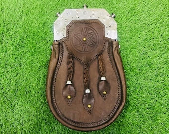 Può includere: Uno sporran in pelle marrone con una parte superiore in metallo argentato decorata con borchie. La patta anteriore presenta un motivo circolare in rilievo con un perno centrale in ottone e tre nappe in pelle pendenti con terminali argentati.